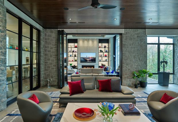 A sophisticated open-plan living room with a large sectional sofa, sculptural lounge chairs, red accent cushions, and a coffee table in the foreground. Rough grey stone walls frame the space, while full-height black-framed glazing and glass partitions bring in natural light. The ceiling is clad in dark timber and includes recessed lighting and a ceiling fan. Built-in display shelving and a television are visible beyond the seating area, with potted plants and contemporary decor adding warmth.