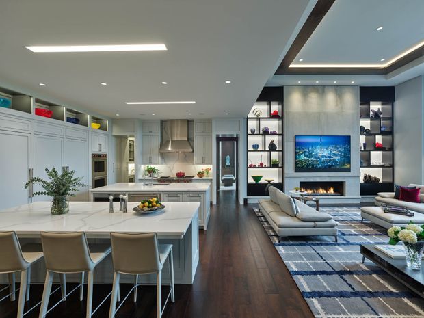 Open-plan contemporary kitchen, dining, and living area with a large white island and integrated cabinetry, dining stools, dark timber flooring, built-in display shelving, a fireplace and television feature wall, recessed lighting, and a neutral sectional seating arrangement. The space is bright, refined, and carefully integrated for entertaining and family living.