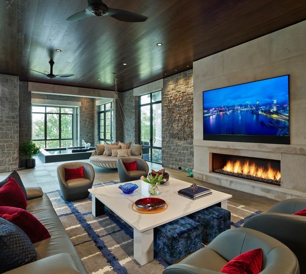 A spacious, sophisticated living room with a large sectional sofa and sculptural lounge chairs arranged around a substantial white coffee table. The room features a dramatic linear fireplace with a wall-mounted television above it, exposed textured stone walls, expansive black-framed glazing, a dark timber-clad ceiling, and contemporary ceiling fans. A patterned area rug and red accent cushions add contrast to the neutral scheme.