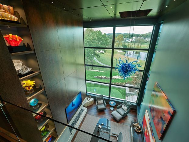 A dramatic double-height living room viewed from above, with floor-to-ceiling glazing overlooking landscaped grounds and a water feature. The room features dark wood wall panelling, built-in display shelving, a television, low coffee table, contemporary lounge seating, colourful artwork, and a striking blue sculptural pendant suspended in front of the glazing.