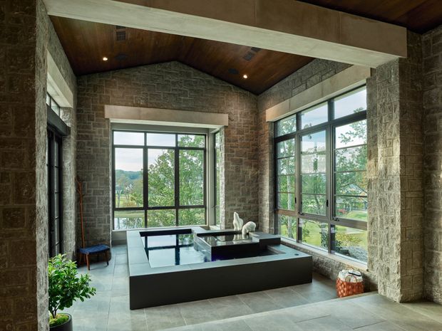 A spacious, rustic-modern living room with exposed stone walls, a pitched timber-lined ceiling, and prominent dark-framed floor-to-ceiling windows overlooking a green landscape. The room contains a large low-profile dark sectional sofa and a central glass-topped coffee table, with a broad structural beam spanning the ceiling.