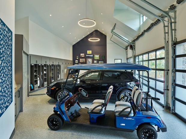 A bright, high-ceilinged garage or showroom-like vehicle storage space with a blue golf cart in the foreground and a dark station wagon behind it. The room has polished concrete or speckled hard flooring, extensive grey built-in locker cabinetry along the left wall, large glazed sectional garage doors on the right, exposed overhead door hardware, recessed ceiling lights, and circular pendant fixtures. A dark feature wall displays framed items and artwork.