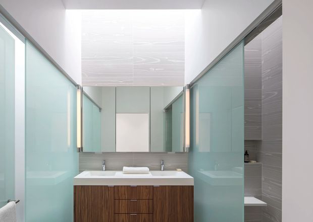 A bright contemporary bathroom vanity area with a double sink, wall-to-wall mirror, and dark timber-look cabinetry. Translucent aqua glass partitions frame the vanity and partially screen a shower or bath zone, while grey textured wall tiles add contrast. The space has clean geometric lines, integrated vertical lighting, and a high, airy ceiling.