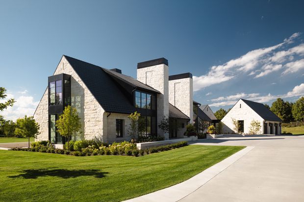 Exterior view of a large residential property with steep dark gable roofs, pale stone masonry walls, tall black-framed glazing, and prominent rectangular stone chimneys. The landscaped grounds feature manicured lawns, planting beds, and a broad concrete driveway leading to a detached garage or outbuilding. The composition has a refined contemporary farmhouse character under clear daylight.