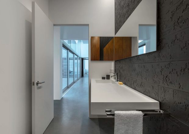 A contemporary bathroom viewed from the doorway, featuring a long white floating vanity with integrated basin, wall-mounted chrome tapware, a large mirror, dark textured feature wall tiles, and warm timber wall cabinetry. A glazed corridor is visible beyond the open door.