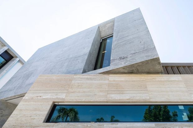 Low-angle exterior view of a contemporary multi-level building facade. The upper volume is formed from smooth, monolithic grey concrete with a narrow vertical window, while the lower facade is clad in pale beige horizontal stone or concrete panels and contains a long horizontal window reflecting trees. A timber-toned slatted element is visible at the roofline on the right.