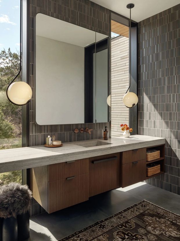 A refined contemporary bathroom vanity with a large rounded-rectangle mirror, twin spherical pendant lights, dark vertically arranged wall tiles, a substantial pale stone benchtop, and warm timber cabinetry. The room has floor-to-ceiling glazing with views to vegetation, a dark floor, open shelving with woven baskets, and a patterned rug.