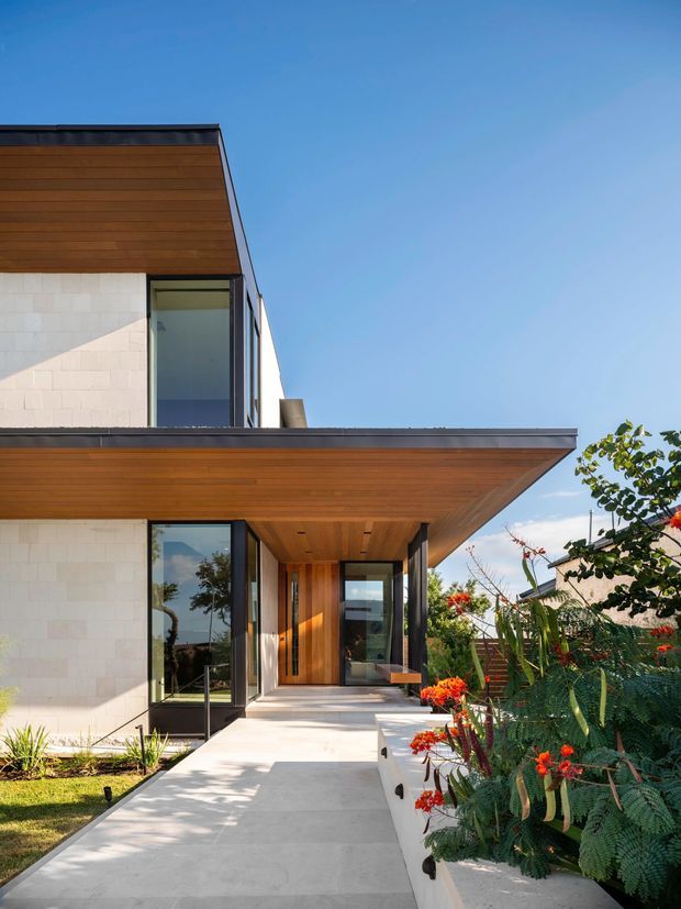 Contemporary residential exterior viewed from the front entry walkway. The house features clean geometric volumes, broad cantilevered roof planes with warm timber soffits, pale masonry wall cladding, dark metal window and door frames, and a timber-lined recessed entry. A landscaped garden borders the concrete path, while strong sunlight creates crisp shadows across the facade.