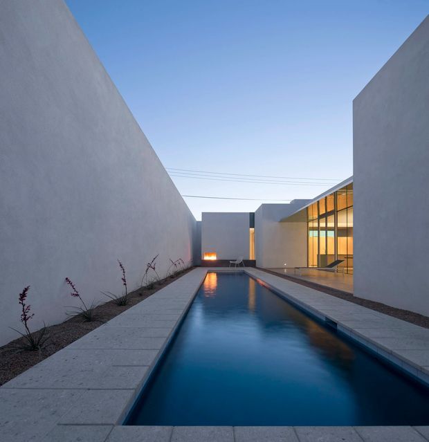 A minimalist architectural courtyard viewed at dusk, centered on a long rectangular swimming pool bordered by large pale paving slabs. Tall smooth rendered walls enclose the space, with sparse ornamental planting along one side. A warm linear fire feature glows at the far end, while a glass-walled interior with timber-toned framing opens onto the pool terrace. The composition is geometric, calm, and highly controlled, with cool blue water contrasting against warm interior lighting.