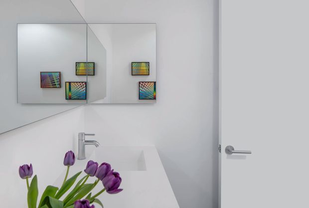 Minimal contemporary bathroom vanity with a white integrated basin and chrome single-lever tap. A large frameless mirrored wall reflects colourful geometric artwork, while a plain white door with a simple chrome lever completes the clean, bright space. Purple tulips provide a small decorative accent in the foreground.