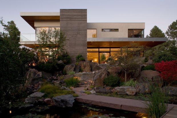 A contemporary multi-level residence viewed from a landscaped garden at dusk. The house has strong horizontal roof planes, broad glazing, projecting cantilevered forms, and a tall central textured concrete or stone-clad volume. Warm interior lighting glows through the lower-level glazing. In the foreground, a naturalistic garden includes large rocks, layered planting, a pond, and a stepping path.