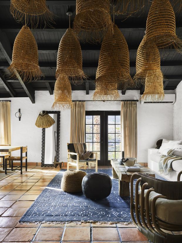 A warmly styled living room with white textured walls, dark exposed ceiling beams, terracotta tiled flooring, and large black-framed French doors. Multiple oversized woven pendant lights hang from the ceiling, complemented by a blue patterned rug, neutral seating, woven lounge chairs, a coffee table, and sculptural ottomans. The space has a relaxed rustic Mediterranean character with strong natural textures and abundant daylight.