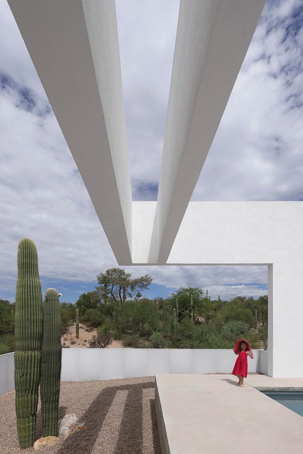 A minimalist desert courtyard or pool terrace framed by dramatic white concrete roof beams and a projecting flat-roof canopy. The scene includes pale concrete paving, a gravel garden with tall columnar cacti and native desert planting, a low white boundary wall, and a partially visible swimming pool. A person in a red dress provides a small contrasting accent. The image strongly emphasizes the architectural canopy against a partly cloudy sky and arid landscape.