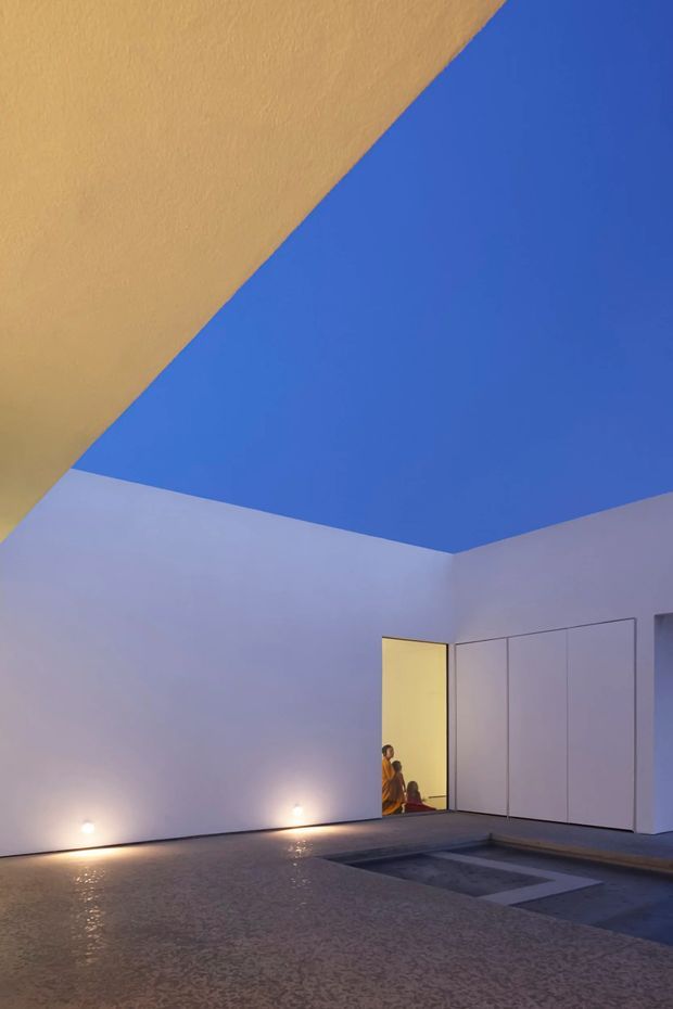 A minimalist architectural courtyard or pool-area scene at dusk, enclosed by smooth white walls and large flush wall panels. The ceiling or overhead plane is painted an intense cobalt blue, contrasting with warm yellow light spilling from an open doorway. Small recessed wall lights illuminate a textured paved surface, with a shallow rectangular water feature or pool set into the foreground.