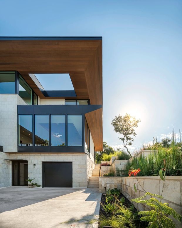 Exterior view of a contemporary multi-level residence with a projecting timber-lined roof canopy, large dark-framed glazed openings, pale stone masonry walls, a garage, and landscaped terraces with steps leading upward. Strong sunlight and clear sky create a bright architectural facade photograph.