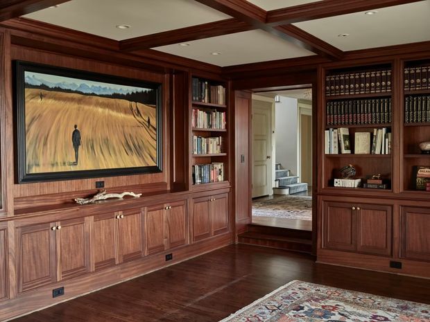 A richly appointed wood-panelled library or study with extensive built-in dark timber cabinetry, bookcases, and lower cupboards. The room has a coffered timber ceiling with recessed lights, polished dark timber flooring, a framed artwork above the cabinetry, and a patterned area rug. An open doorway leads to a hallway and stair area beyond.