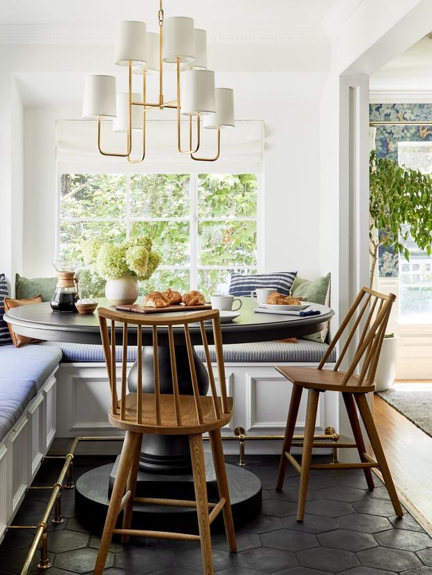 Bright, refined breakfast nook or dining area with a built-in curved banquette surrounding a round dark dining table. The space features white panelled walls, large divided windows, a brass multi-arm pendant with fabric shades, mid-century-inspired wooden dining chairs, patterned blue cushions and drapery, and dark hexagonal floor tiles. A small adjacent area with leafy patterned wallpaper is visible through the opening on the right.
