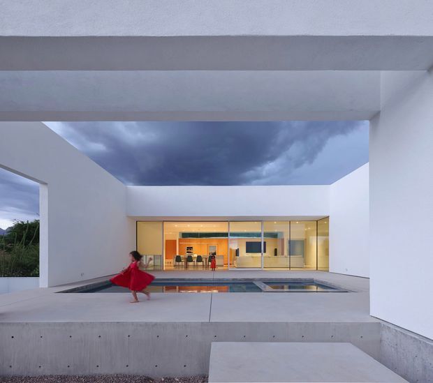 A minimalist modern courtyard viewed from an open covered terrace, framed by crisp white and pale concrete walls. A shallow reflecting pool occupies the foreground, with a glazed, warmly lit interior pavilion beyond. The composition emphasizes strong horizontal and rectangular architectural forms, open sky, and a dramatic cloudy atmosphere.