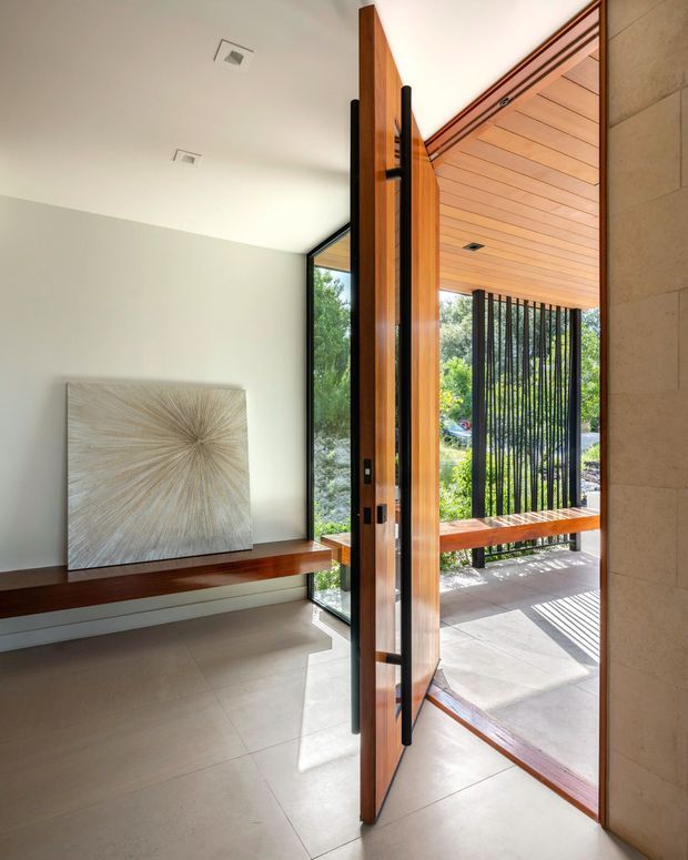 Bright contemporary entryway viewed from inside, featuring a dramatic full-height timber pivot door with black hardware, pale tiled flooring, white walls, and a built-in dark timber bench with artwork. The open door reveals a covered outdoor porch with a timber-lined ceiling, vertical black screening, and abundant greenery beyond.