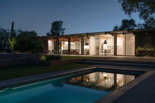 Twilight exterior view of a low, single-storey modern home opening onto a broad terrace and rectangular swimming pool. The house has extensive floor-to-ceiling glazing, slender dark frames, a long shallow roofline, warm interior lighting, and a restrained flat-roofed composition. The surrounding landscaped garden and pool create a calm indoor-outdoor setting.