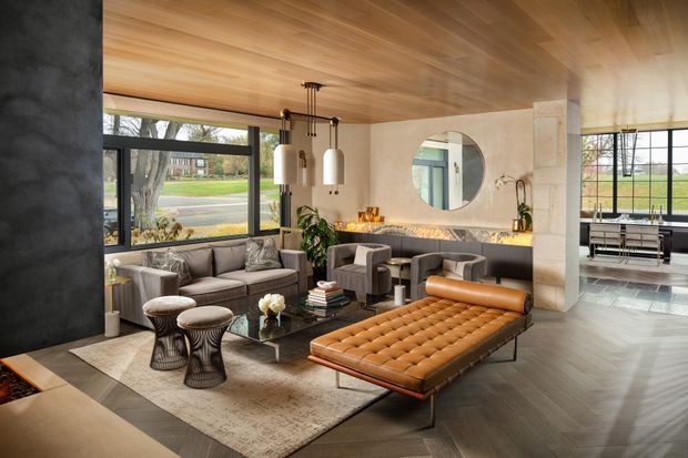 A spacious, refined open-plan living room with a low tufted leather chaise lounge, grey upholstered sofas and armchairs, sculptural side tables, and a large neutral rug. The room features extensive black-framed glazing, a dark textured feature wall, a warm timber-lined ceiling, a stone or plaster fireplace wall with a round mirror, and a dining area visible beyond.