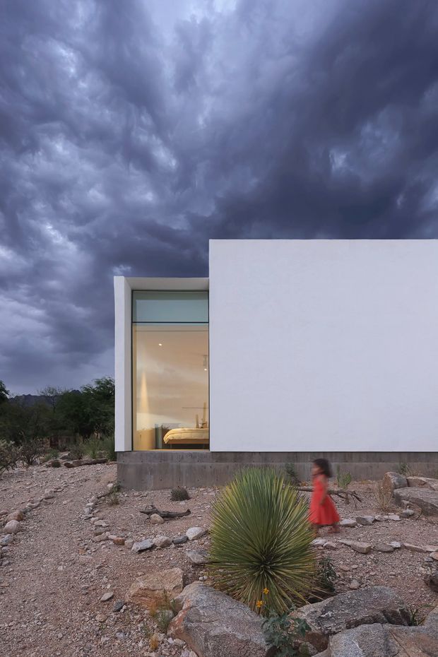 A minimalist single-storey white geometric house set in a rocky, arid landscape beneath dramatic storm clouds. The facade features a large floor-to-ceiling glazed opening with a visible bedroom inside, alongside a broad blank white wall and exposed concrete base. Native drought-tolerant planting and boulders form the sparse landscape in the foreground.