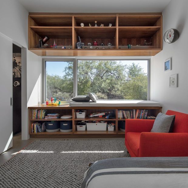 Bright, compact reading and family room with extensive custom walnut-toned built-in shelving around a large picture window. The lower joinery provides open book storage and baskets, while a red upholstered armchair and grey throw-covered sofa frame a textured grey rug. White walls, abundant daylight, and views of trees create a warm, practical, inviting space.