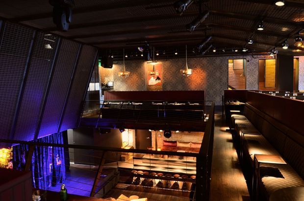 A dark, dramatic multi-level hospitality interior, likely a bar or restaurant, viewed from an upper level. The space features extensive black metal mesh screens, dark timber or stained wood seating and bar joinery, patterned wall finishes, pendant lights, theatrical spotlights, and vivid blue and amber accent lighting. Long upholstered banquette seating lines the right side, with additional dining or bar seating visible below around an open void.
