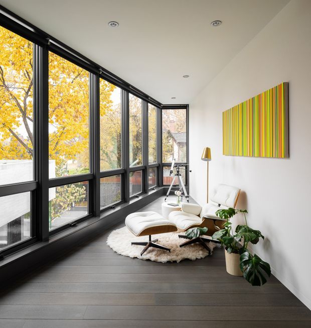 A bright, elongated sunroom or enclosed lounge with full-height black-framed glazing along one side, overlooking autumn trees. The room has dark timber-look flooring, white walls and ceiling with recessed downlights, a white leather lounge chair and ottoman, a sculptural floor lamp, a tripod-style telescope, indoor plants, a shaggy cream rug, and a colourful vertically striped wall artwork. The space feels airy, contemporary, and inviting.