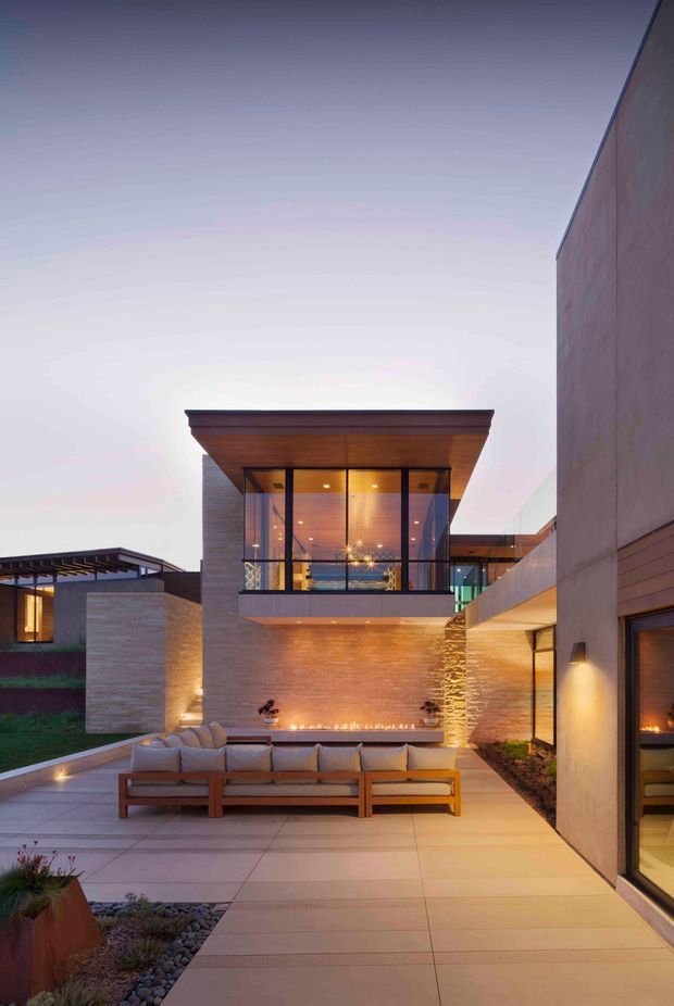Twilight view of a refined contemporary residential courtyard and outdoor entertaining area. Large-format paving surrounds a timber-framed outdoor lounge with cushioned seating, a linear fire feature set into a warm stone-clad wall, and extensive floor-to-ceiling glazing. The multi-level house has clean rectilinear forms, dark-framed glass, timber soffits, and integrated landscape planting.