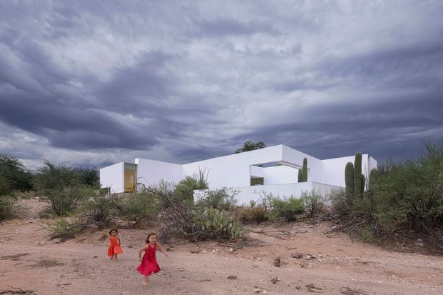 Exterior view of a stark white, low-profile modern house set in a sparse desert landscape, with flat roof planes, rectilinear forms, minimal openings, and tall columnar cacti. Two children are visible in the foreground beneath a dramatic overcast sky.