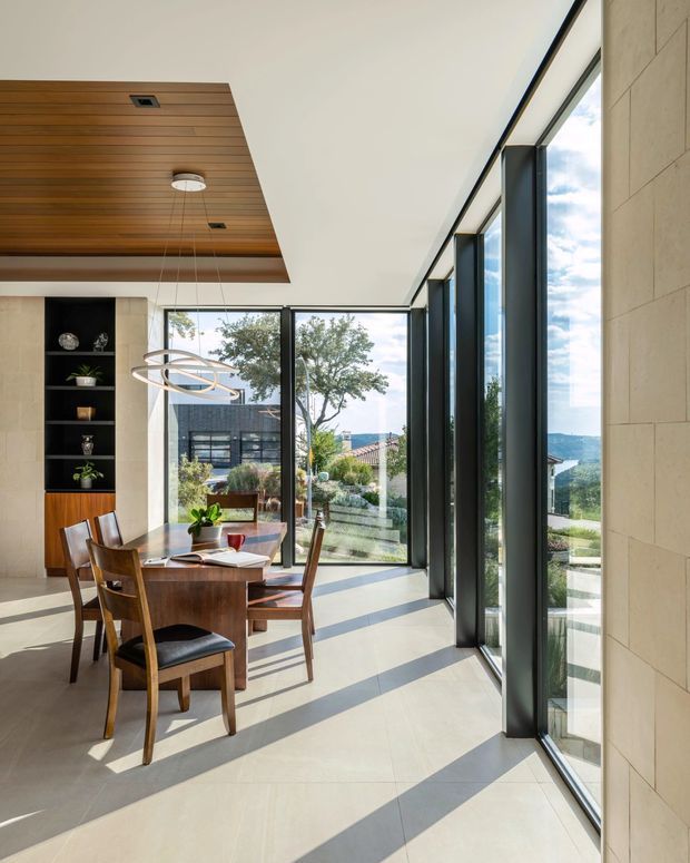 A bright, modern dining room with a timber dining table and matching wooden chairs beside extensive floor-to-ceiling glazing. The room features a warm timber-paneled ceiling inset, a sculptural contemporary pendant light, a dark built-in display shelving unit, pale large-format flooring, and light stone or masonry wall surfaces. Strong afternoon sunlight casts long shadows across the floor, with landscaped residential surroundings visible outside.