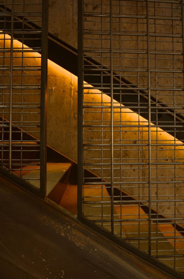 Close-up architectural detail of an industrial stairway enclosed by a rigid steel grid or mesh screen. Warm amber lighting illuminates timber or plywood stair surfaces behind the dark metal framework, creating a dramatic, utilitarian interior composition.