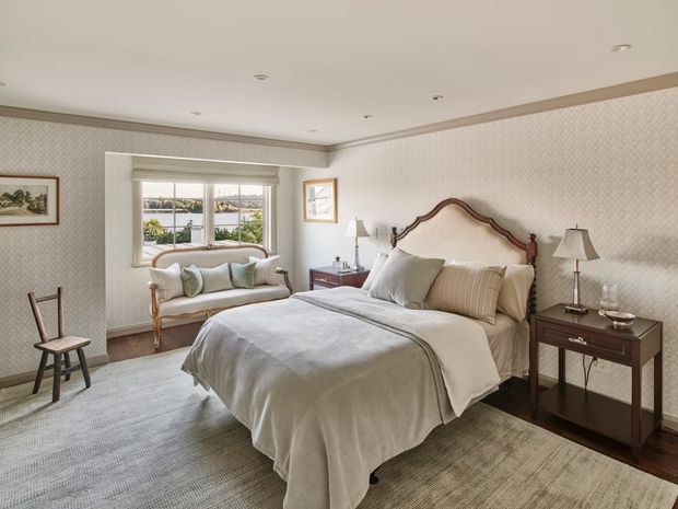 A traditional bedroom with a carved wooden sleigh-style bed, upholstered bedding, matching bedside tables and lamps, a small settee beneath a window, patterned wallpaper, dark timber flooring, and a large neutral rug. The room has soft natural light and a calm, refined atmosphere.