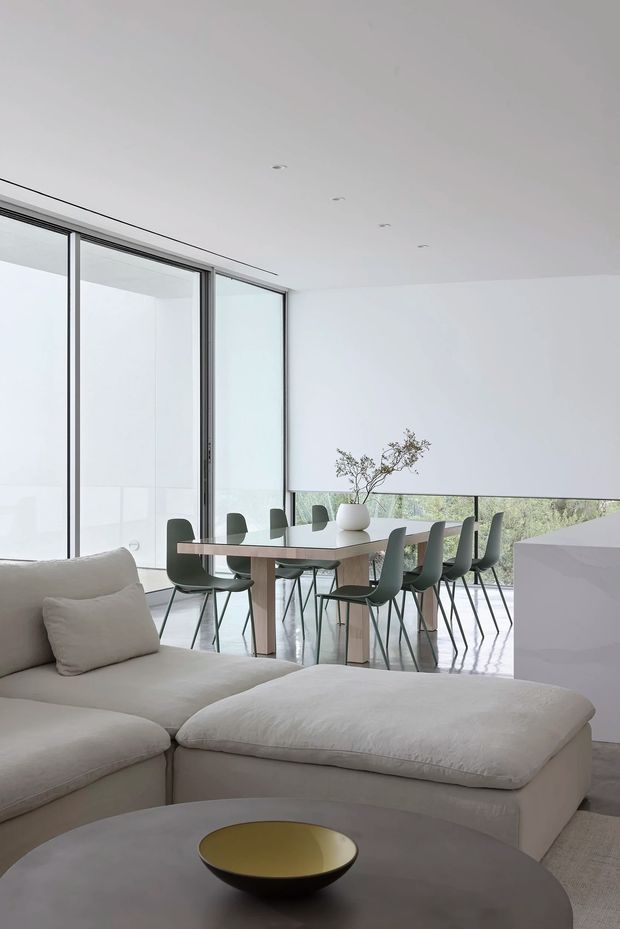 A bright, minimalist open-plan interior with a white living area in the foreground and a dining space behind it. A large cream sectional sofa surrounds a round dark coffee table with a small bowl, while a timber dining table is paired with multiple dark green upholstered chairs. Full-height sliding glass doors and a glazed balustrade/opening bring in views of greenery. The room has a smooth white ceiling with recessed downlights, pale reflective flooring, and restrained contemporary detailing.