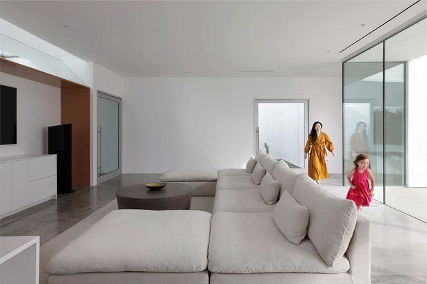 Bright, minimalist contemporary living room with a large off-white sectional sofa and ottoman, a dark round coffee table, white built-in media cabinetry, and expansive floor-to-ceiling glass doors. The room has a clean open-plan feel with polished grey flooring and strong natural light.