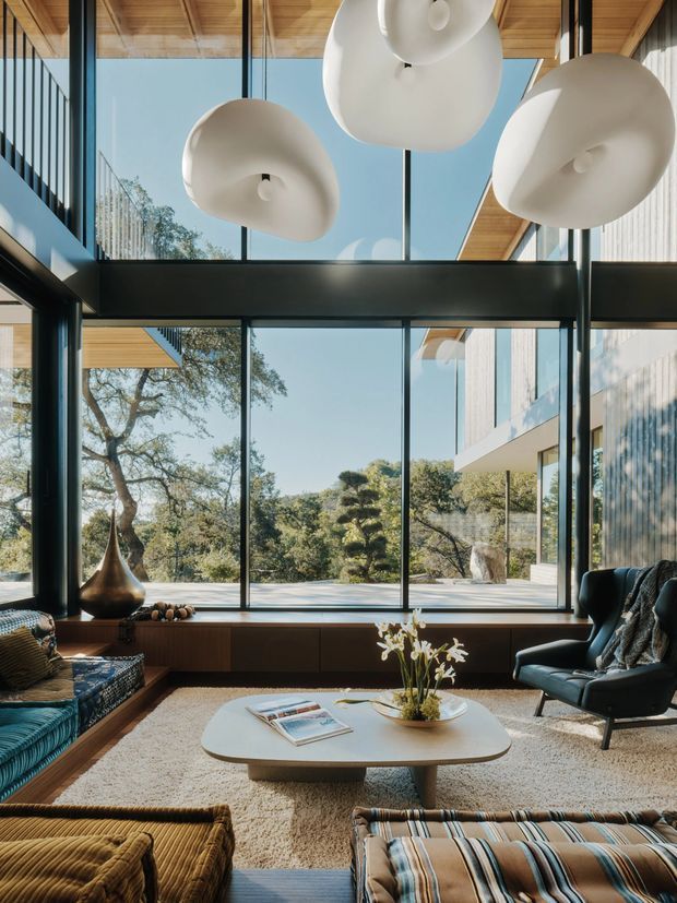 A spacious mid-century-inspired living room with floor-to-ceiling black-framed glazing, warm timber ceiling panels and built-in timber cabinetry. Sculptural white pendant lights hang above a low oval coffee table, while upholstered lounge seating and cushions create a relaxed, curated setting. Trees and a landscaped exterior are visible through the extensive glazing.