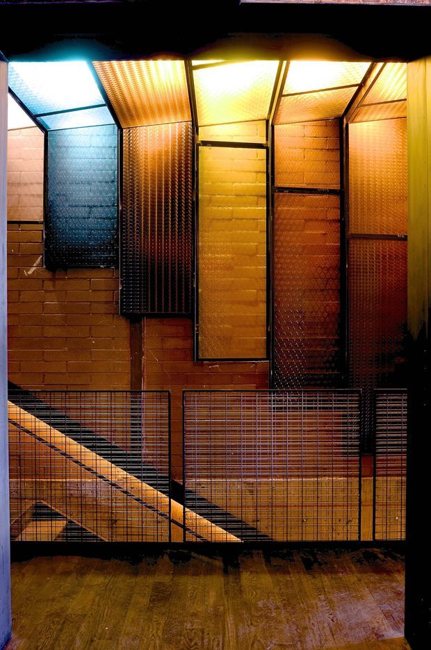 Interior view of a dramatic industrial-style stairwell or landing, featuring exposed reddish-brown brick walls, black metal framing, wire-mesh balustrades, and translucent corrugated or textured panels illuminated with strong amber and blue light. A timber-toned floor is visible in the foreground, with a staircase descending behind the mesh railing.