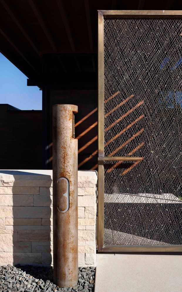 Close-up exterior architectural detail showing a rusted cylindrical metal post beside a large framed diamond-mesh metal screen or gate. A pale rendered or masonry block wall, dark timber roof structure, and strong diagonal sunlight shadows are visible. The composition emphasizes raw industrial materials, weathering, and layered screening.