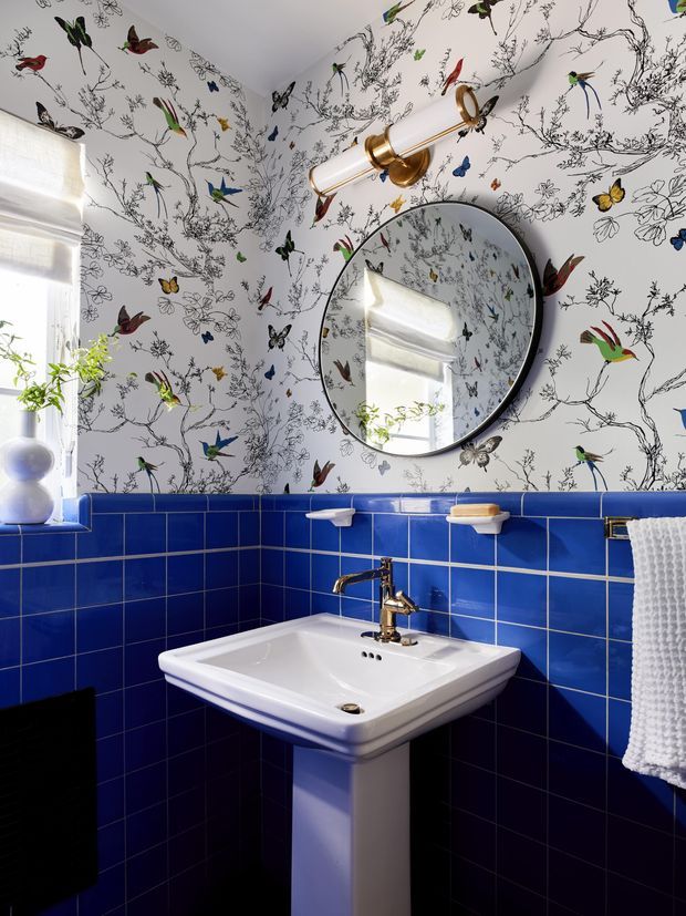 A bright, compact bathroom with a pedestal basin beneath a round mirror. The lower walls are clad in glossy cobalt-blue square tiles, while the upper walls feature lively white wallpaper decorated with illustrated birds, butterflies and branches. A brass wall light, brass tapware and white textured towel add decorative detail. Natural light enters through the window at left.