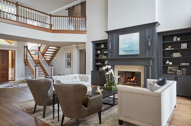 A refined double-height living room with a prominent dark painted fireplace and built-in shelving wall, displaying artwork and decor. The room includes sofas, upholstered armchairs, a coffee table, a patterned area rug, timber flooring, and a timber staircase with white balustrades leading to an upper landing. Abundant glazing and an open mezzanine create a bright, spacious interior.