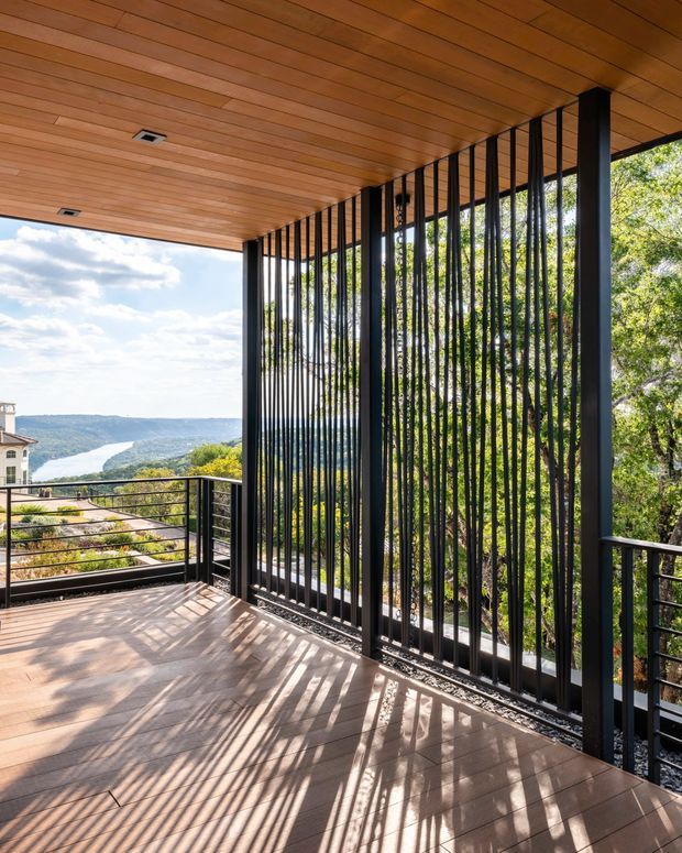 A sunlit covered outdoor terrace or balcony with warm timber decking and a matching timber-lined ceiling. Tall black vertical metal screens form a balustrade and privacy feature along the edge, casting strong linear shadows across the floor. A black horizontal railing and a scenic tree-covered valley with a winding body of water are visible beyond the open side. The space has a contemporary architectural character with a strong indoor-outdoor connection.