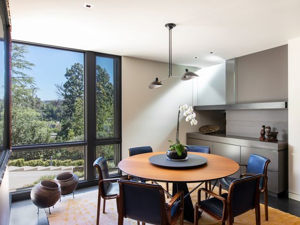 A refined dining room with a round timber dining table and six dark upholstered chairs, positioned beside large floor-to-ceiling windows overlooking leafy surroundings. The room features dark-framed glazing, a warm neutral wall, a built-in dark wood or dark-toned display and storage unit, contemporary pendant lighting, and a patterned rug. The overall design is polished and mid-century influenced, with abundant natural light.