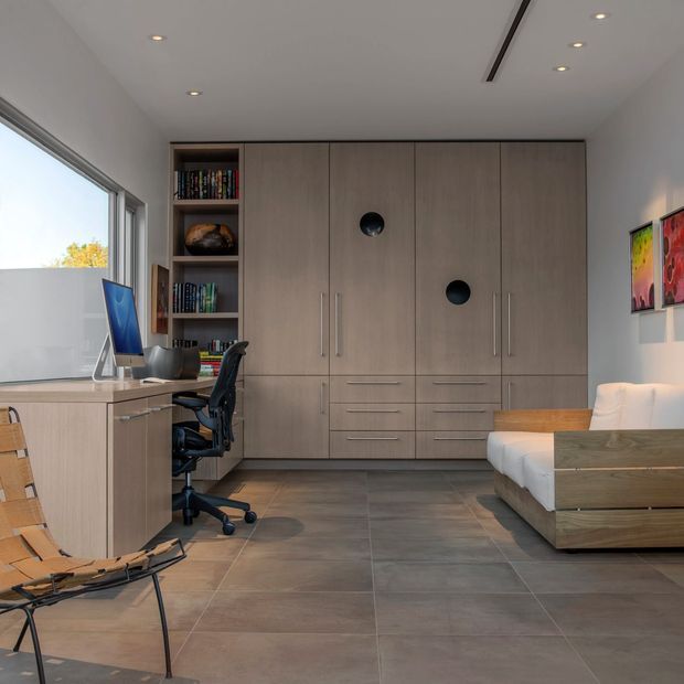 A compact, highly organized multipurpose room with a built-in timber-toned wall of wardrobes and drawers, integrated open shelving, and a dedicated desk workspace. The room also includes a modern lounge chair, a low timber bench or daybed with cushions, large-format grey floor tiles, recessed ceiling lights, and broad glazing along the left side. The palette is restrained and neutral, with warm wood cabinetry, grey flooring, white walls, and small colourful artworks.
