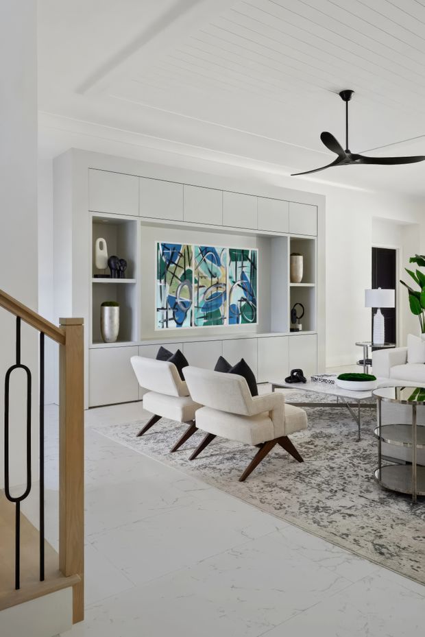 Bright, refined living room with a full-width built-in white media and display wall, abstract artwork, sculptural objects, two cream lounge chairs, a pale patterned rug, marble-look flooring, and a sleek black ceiling fan. A timber-and-black-metal stair balustrade is visible in the foreground, with contemporary furniture and restrained monochromatic styling throughout.