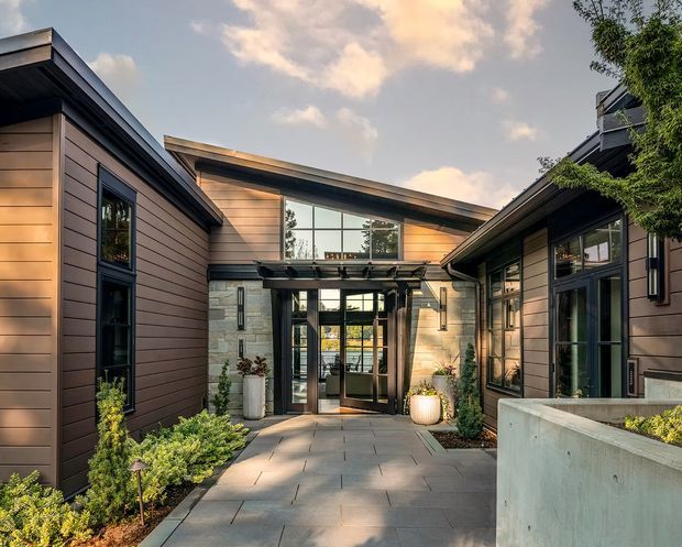 A contemporary residential entry courtyard viewed from outside, with a paved walkway leading to a glazed front door beneath a projecting canopy. The building has low-pitched roof forms, dark metal window and door frames, horizontal timber-look cladding, pale masonry feature walls, and integrated landscaping in raised planters. Warm late-day light creates a refined, welcoming exterior composition.