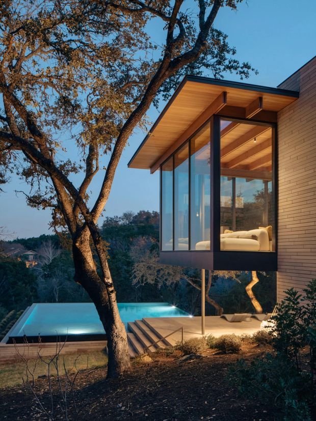 A contemporary multi-level house set among mature trees on a sloping landscaped site, photographed at dusk. The building features a projecting glazed room with full-height windows, a broad flat roof overhang, warm timber-lined soffits and ceilings, and a pale brick-clad wall. A lit swimming pool and stepped outdoor terrace are visible below, with a softly furnished interior seen through the glazing.