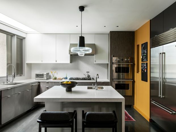 Contemporary kitchen featuring a large white island with two black bar stools, dark lower cabinetry, white upper cabinets, integrated stainless-steel appliances, and a warm mustard-yellow accent wall. A large window, pendant light, timber-toned pantry cabinetry, and dark flooring complete the clean, high-contrast scheme.