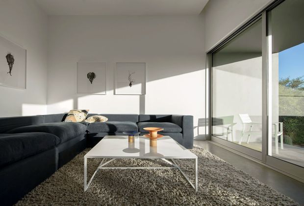 A bright contemporary living room with a large dark charcoal sectional sofa arranged around a white rectangular coffee table. A textured grey rug, sculptural small tables, and minimalist framed artworks furnish the room. Full-height sliding glass doors open to an outdoor terrace, bringing in strong sunlight and views of greenery. The space has white walls, a simple flat ceiling, and a calm, minimalist aesthetic.