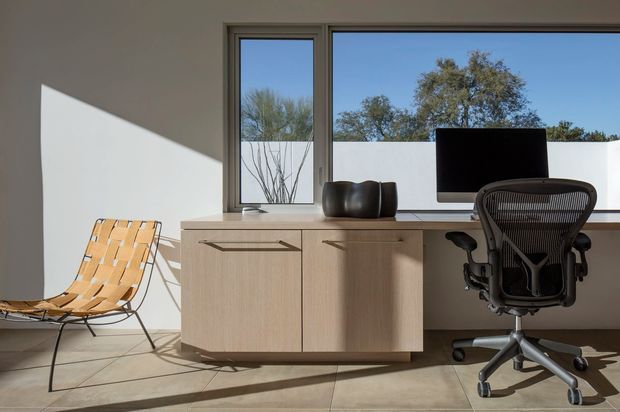 A minimalist home office with a pale wood built-in credenza beneath a wide window, a black monitor and ergonomic office chair, and a woven lounge chair. Strong sunlight creates angular shadows across the white walls and smooth floor, with trees visible outside.